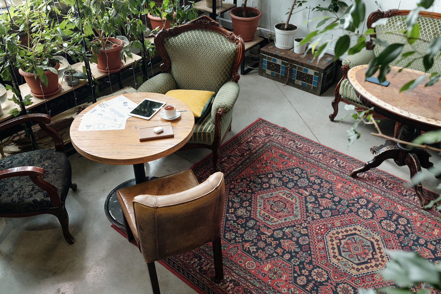 A cozy café corner with vintage chairs, a round table holding a tablet, documents, and a drink, surrounded by potted plants and a patterned rug on the floor.