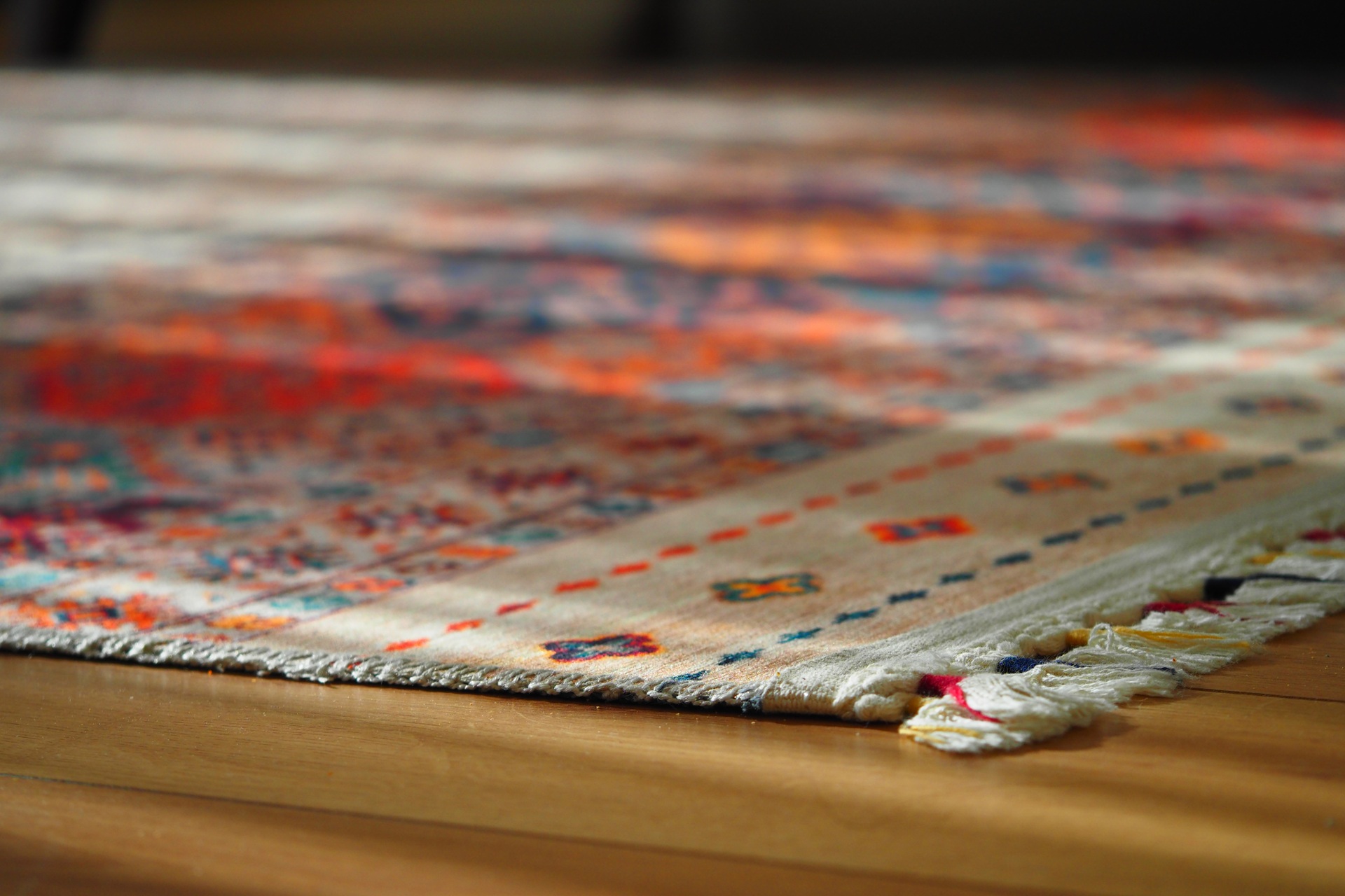 Close-up of the edge of a patterned rug with fringed ends, lying on a wooden floor, showing colorful designs and detailed stitching.