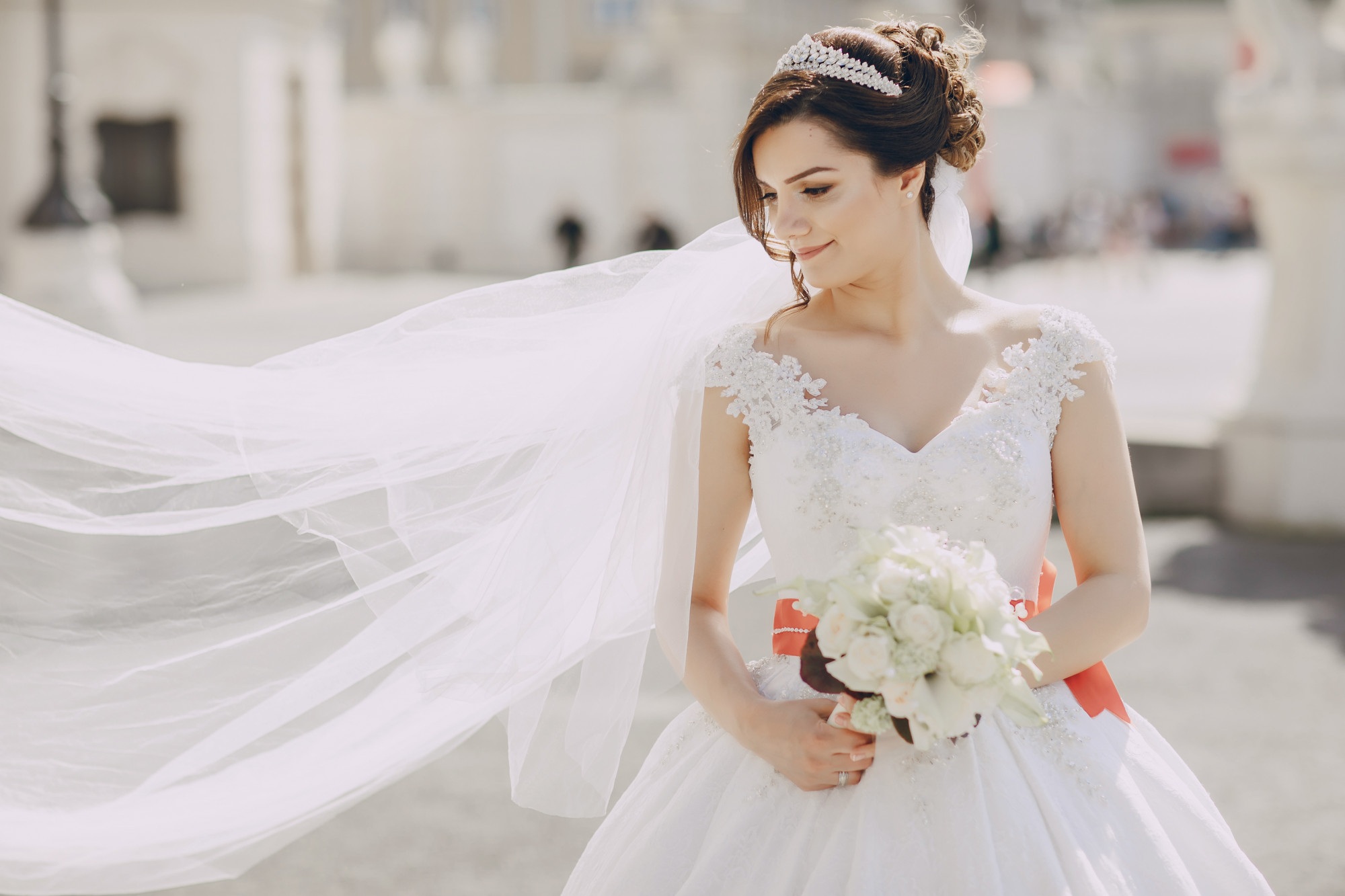 Bride in a white wedding dress with lace details and a tiara holds a bouquet of white flowers; her veil is blowing in the wind outdoors.