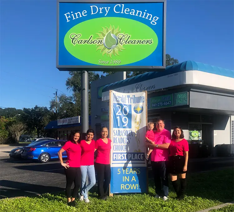 Six people in matching pink shirts stand outside Carlson Cleaners, holding a banner for a 2019 first place award in front of a dry cleaning business sign.
