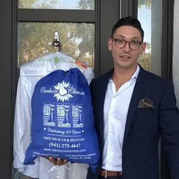 A man in a suit holds a blue Carlson Cleaners bag in front of a door with clothing in plastic on a hanger behind him.