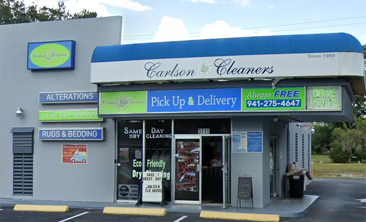 A storefront for Carlson Cleaners, featuring signs for alterations, same day service, rugs and bedding, pick up and delivery, and a drive-thru, with contact info displayed.