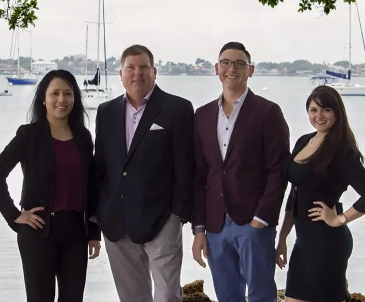 Four people dressed in business attire stand outdoors in front of a body of water with sailboats and buildings in the background.
