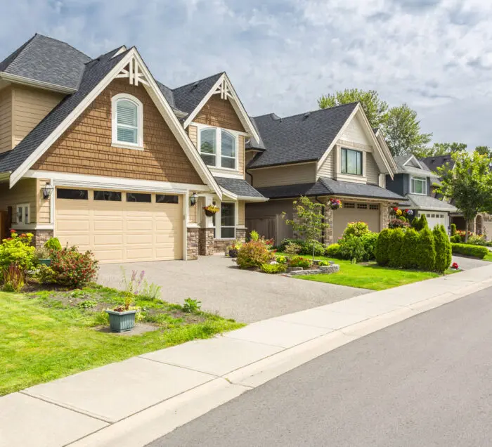 Row of modern suburban houses with attached garages, well-maintained front lawns, and landscaping along a residential street under a partly cloudy sky.