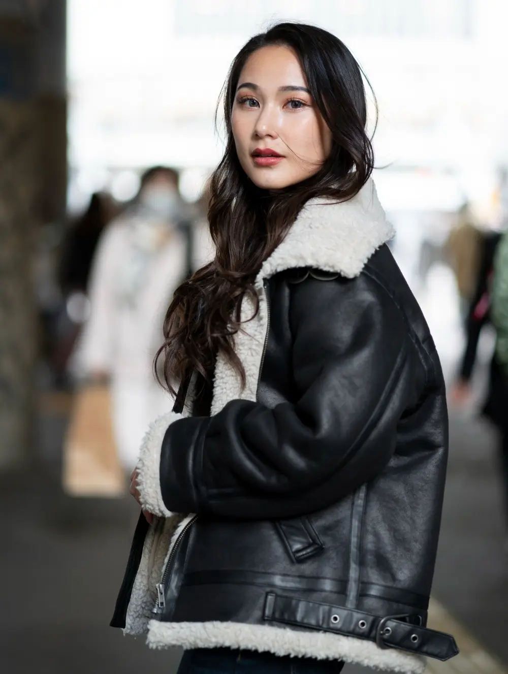 A woman with long dark hair wears a black shearling jacket, standing outdoors with blurred people and buildings in the background.