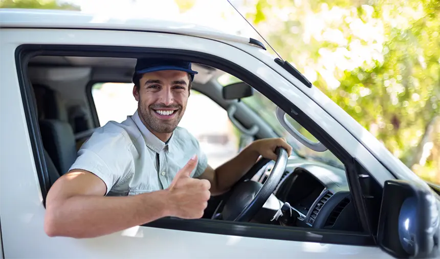 A man in a light shirt and blue cap sits in the driver’s seat of a white vehicle, smiling and giving a thumbs-up out the window.