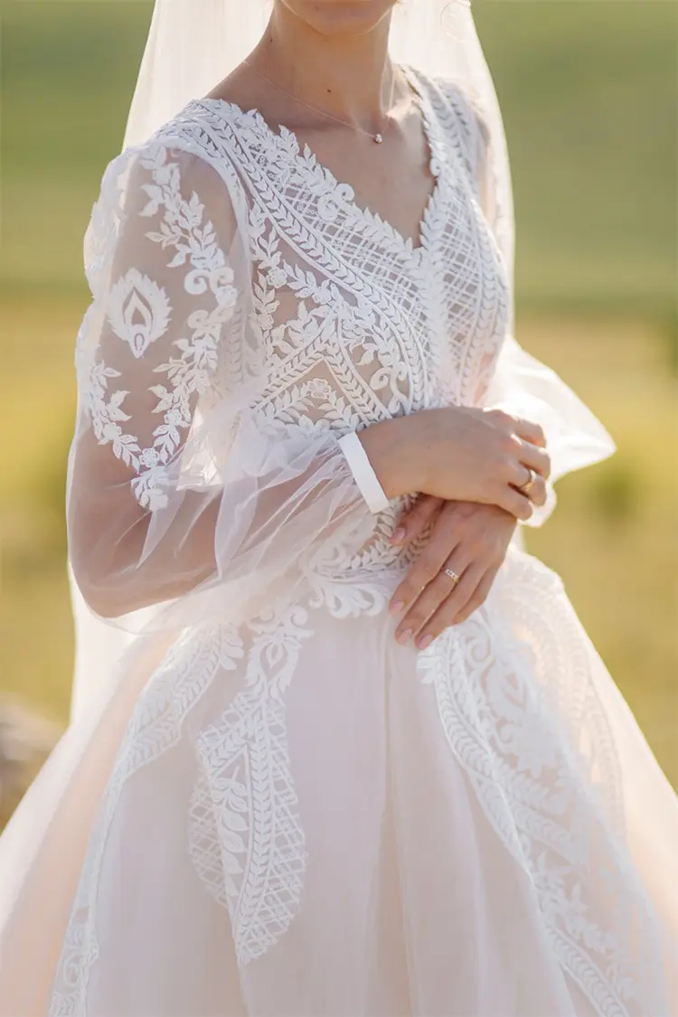 A bride wearing a white lace wedding dress with sheer long sleeves and intricate embroidery, standing outdoors.